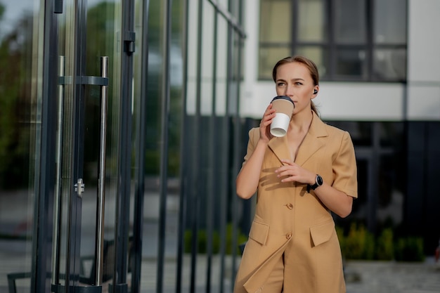 Schöne Frau, die mit Kaffee in der Nähe des Bürogebäudes zur Arbeit geht. Porträt der erfolgreichen Geschäftsfrau, die Tasse heißes Getränk in der Hand auf ihrem Weg zur Arbeit auf der Stadtstraße hält. Hohe Auflösung.