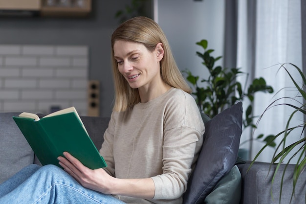 Schöne Frau, die sich nach der Arbeit zu Hause entspannt und ein interessantes Buch liest, das auf der Couch sitzt