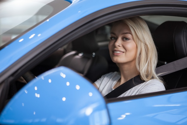 Schöne Frau, die sicher in ihrem Neuwagen hinter dem Lenkrad lächelt zur Kamera sitzt