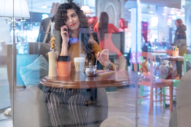 Schöne Frau, die telefoniert, während sie an einem Tisch in einem Café hinter dem Glas sitzt