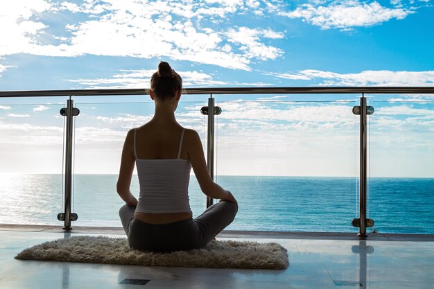 Schöne Frau, die Yoga auf einer Gymnastikmatte praktiziert und Assana-Pose auf einem Balkon mit Meerblick sitzt