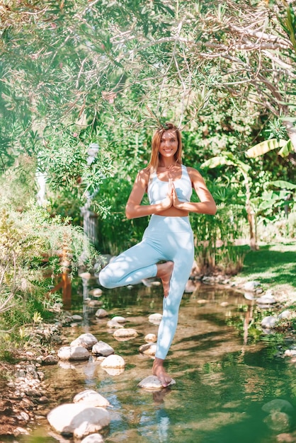 Schöne Frau, die Yoga in einer Baumposition praktiziert Sie steht auf dem Stein im Fluss zwischen Bäumen