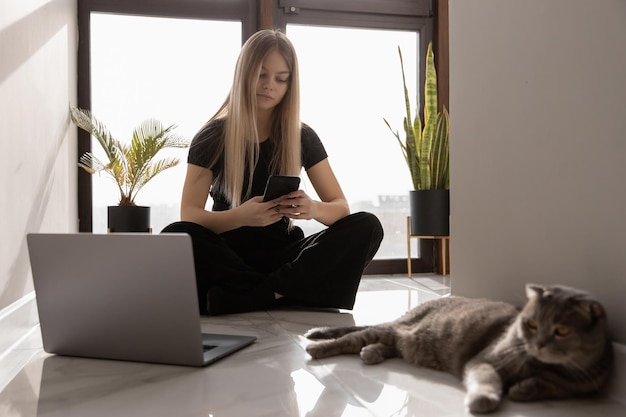 Schöne Frau, die zu Hause am Computer arbeitet, am Fenster auf dem Boden sitzt und eine Nachricht am Telefon eingibt