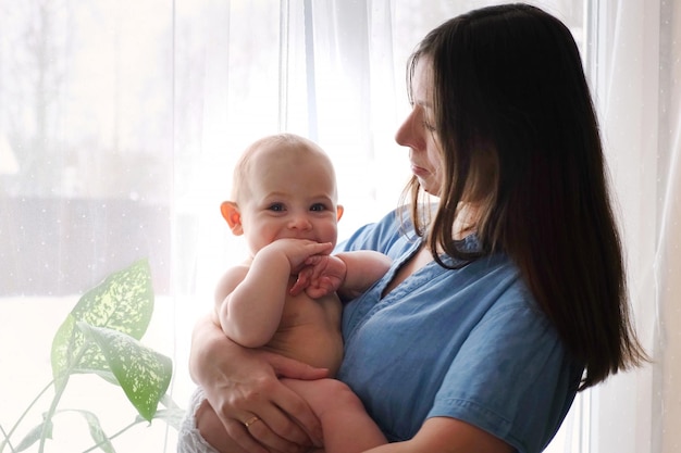 Schöne Frau hält ein neugeborenes Baby Liebevolle Mutter, die ihr Baby zu Hause umarmt Helles Porträt einer glücklichen Mutter, die ein Kind an den Händen hält Liebevolle Mama, die für ein Fenster bleibt und ein entzückendes Mädchen beruhigt