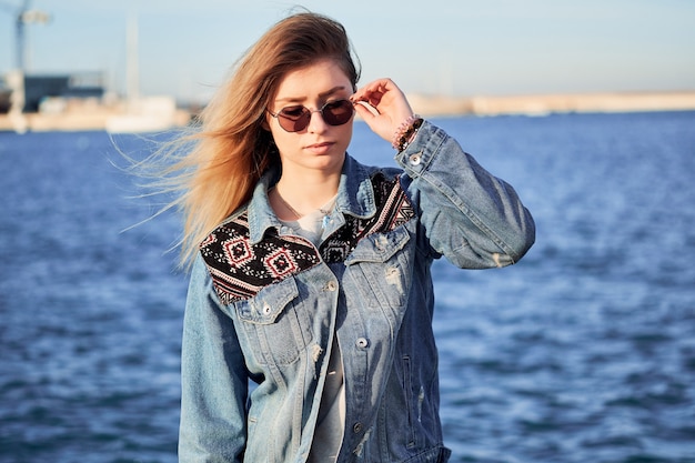 Schöne Frau hält runde Brille in einer Jeansjacke mit langen blonden Haaren an einem Sommertag im Freien im Hafen