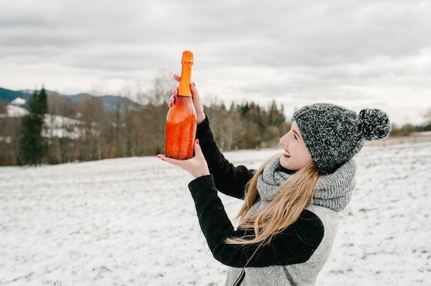 Schöne Frau halten eine Flasche Champagner vor dem Hintergrund der Winterberge. Mädchen im verschneiten Winter, Spaziergang in der Natur. Reise- und Urlaubskonzept. Feiertage Wintersaison.