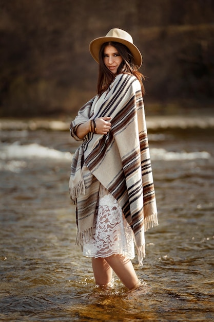 Schöne Frau Hipster mit Hut und Poncho stehen im Wasser des Flusses stilvolles Outfit Boho Reisekonzept