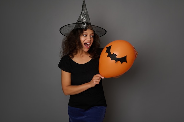 Schöne Frau im schwarzen Hexenkostüm des Karnevals wirft mit einem bunten orange Luftball auf grauem Hintergrund mit Kopienraum auf. Halloween-Konzept für Werbung und Promotion.