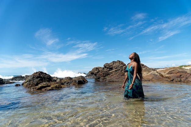 Schöne Frau in blauen Kleidern und Zöpfen im Haar steht am Strand, geht im Wasser und schaut auf die Sonne