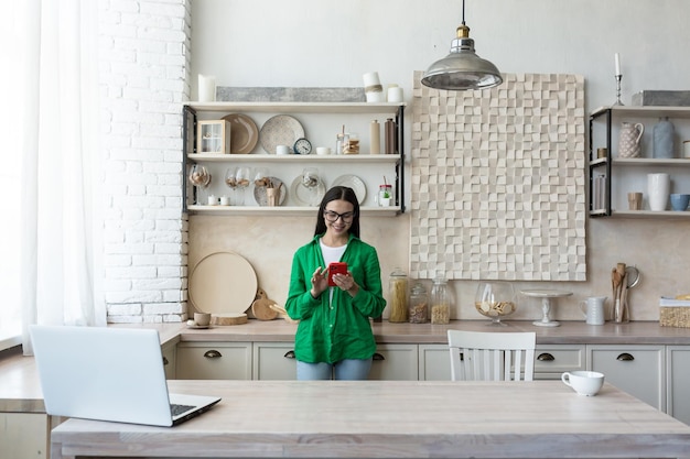 Schöne Frau in Brille und grünem Hemd mit rotem Handy zu Hause in der Küche lächelnd und