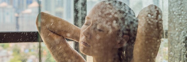 Schöne Frau in der Dusche hinter Glas mit Tropfen auf dem Hintergrund eines Fensters mit Panoramablick auf die Stadt BANNER im Langformat