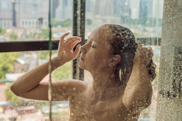 Schöne Frau in der Dusche hinter Glas mit Tropfen auf dem Hintergrund eines Fensters mit Panoramablick auf die Stadt