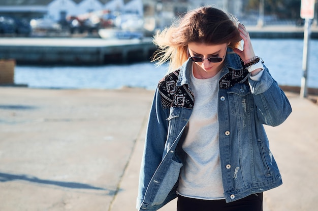 Schöne Frau in der runden Sonnenbrille und in einer Jeansjacke, die langes blondes Haar an einem Sommertag draußen im Hafen nach unten schaut