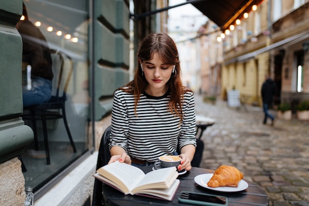 Schöne Frau in drahtlosen Kopfhörern, die ein Buch auf der Café-Terrasse mit frischem Kaffee und Croissant liest