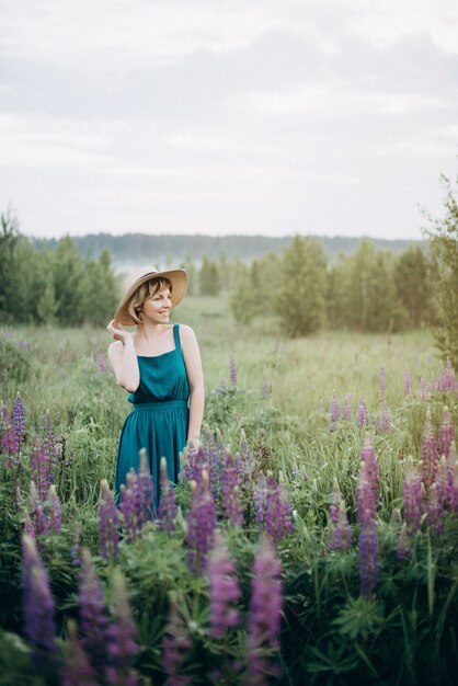 Schöne Frau in einem blauen Kleid in einem Feld von Lupinenblumen und einem Hut bei Sonnenaufgang.