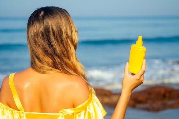 Schöne Frau in einem gelben Seidenkleid besprüht ein Sonnenspray am Strand. Konzept von SPF perfekte Haut rote Lippen helles Make-up Tropen Sommer am Strand