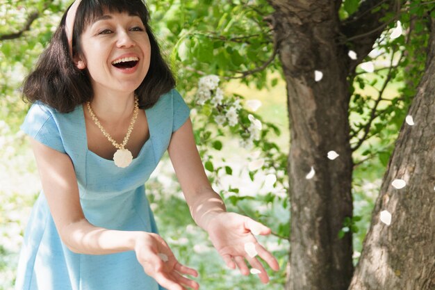 Schöne Frau in einem langen blauen Kleid mit blühenden Apfelbäumen im Frühlingsgarten, sie wirft Blütenblätter