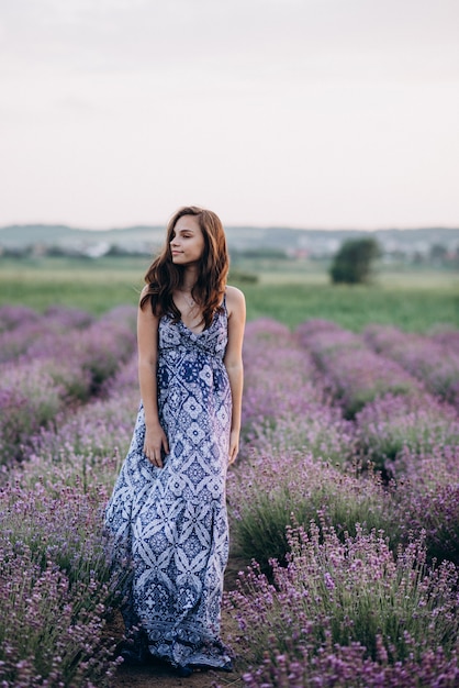 schöne Frau in einem langen Kleid, das in einem Lavendelfeld bei Sonnenuntergang geht.