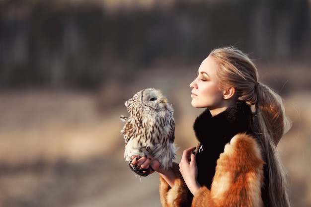 Schöne Frau in einem Pelzmantel mit einer Eule auf seinem Arm. Blondine mit dem langen Haar in der Natur, die eine Eule hält. Romantisches empfindliches Bild einer Frau