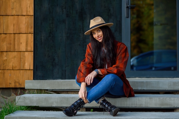 Schöne Frau in einem roten Hemd, Jeans und Hut sitzt auf der Treppe ihres modernen Hauses in der Nähe des Waldes.