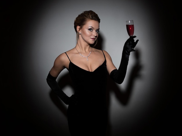 Schöne Frau in einem schwarzen Kleid mit Ausschnitt und langen Samthandschuhen, die tanzen und Champagner halten ...