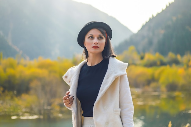Schöne Frau in einem weißen Mantel und schwarzem Hut, die auf einem Hintergrund der Herbstberge aufwirft