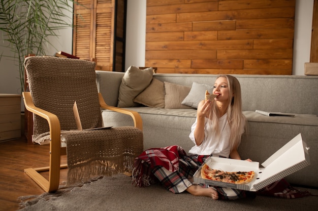 Schöne Frau in einem weißen T-Shirt isst Pizza mit Vergnügen, während sie auf dem Boden neben dem Sofa sitzt