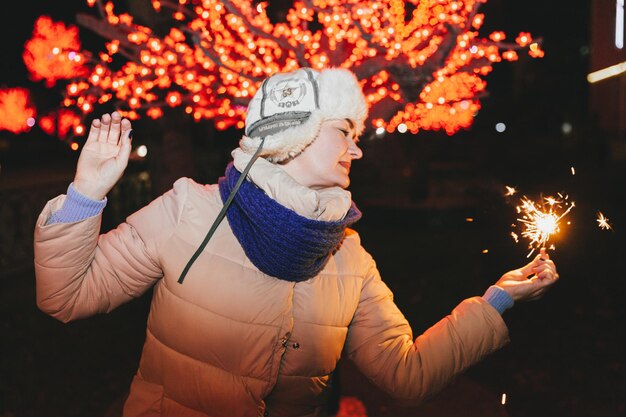 Schöne Frau in einer Strickmütze und einem Schal, die mit einer Wunderkerze in der Stadt stehen. Feier- und Weihnachtskonzept.