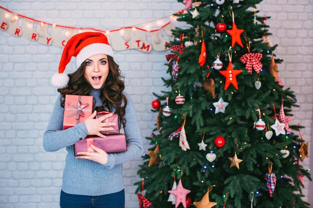 Schöne Frau in einer Weihnachtsmütze mit vielen Geschenkboxen auf Weihnachtsbaum. Neujahr und Weihnachten feiern.