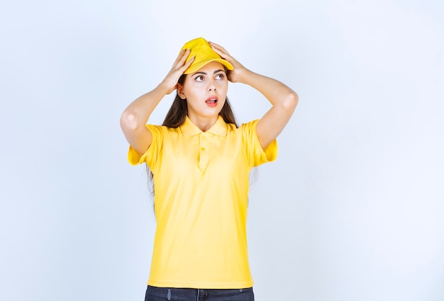 Schöne Frau in gelbem T-Shirt und Mütze schockiert über etwas auf weißem Hintergrund.