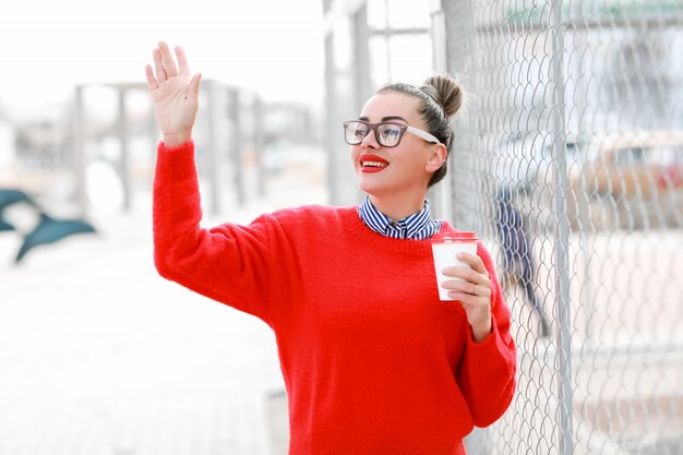 Schöne Frau in Gläsern, rotem Pullover und rotem Lippenstift, die die Straße mit einem Glas Kaffee entlang gehen