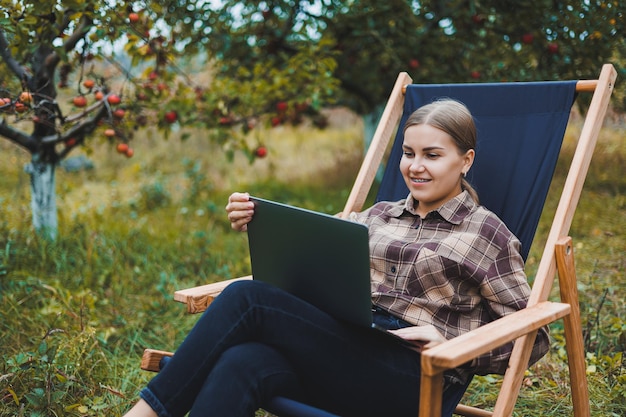 Schöne Frau in kariertem Hemd mit Laptop, die draußen im Garten-Home-Office-Konzept arbeitet Fernarbeit