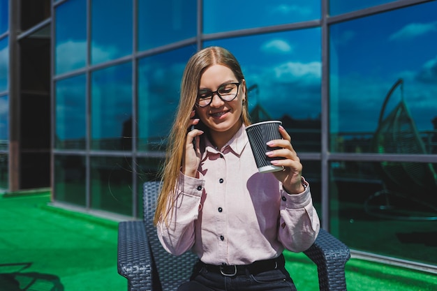 Schöne Frau in klassischer Brille und trendigem Outfit, die eine Nachricht auf einem Handy liest, das neben einem modernen Glasgebäude steht