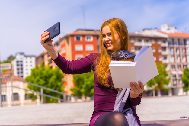 Schöne Frau in lila Kleid und Schal, die ein Buch in einem Park in der Stadt liest und ein Selfie mit dem Handy macht