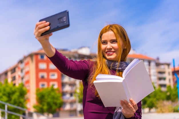 Schöne Frau in lila Kleid und Schal mit einem Buch in der Hand in einem Park in der Stadt, ein Selfie mit dem Handy machen