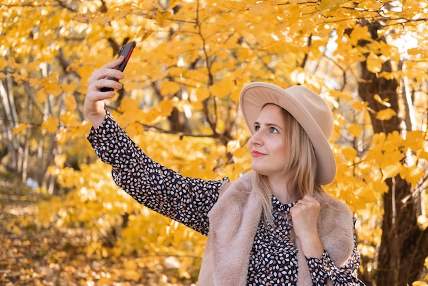 Schöne Frau in modischer Kleidung und Hut macht Selfie am Telefon in der Nähe des gelben Herbstbaums.