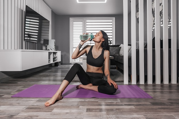 Schöne Frau in Sportkleidung sitzt auf einer Yogamatte und trinkt Wasser