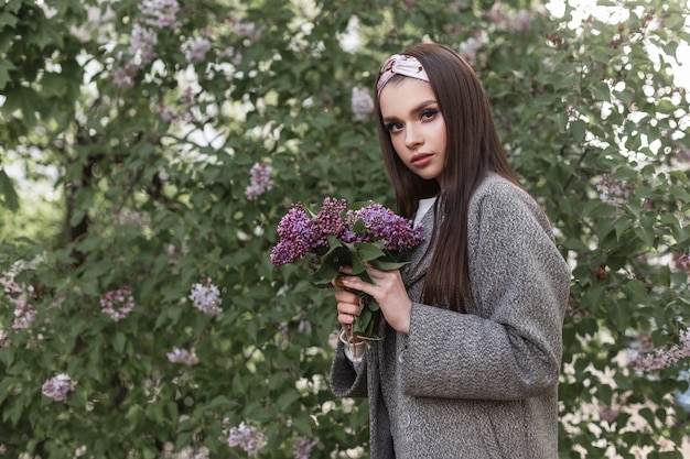 Schöne Frau in stilvollem Bandana in modischer Frühlingskleidung mit Blumenstrauß in den Händen ruht in der Nähe eines blühenden erstaunlichen Baumes am Frühlingstag auf der Straße. Attraktives Mädchenmode-Modell mit purpurroten Blumen.