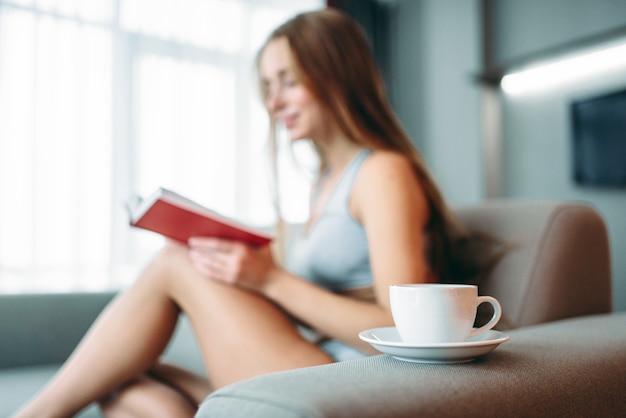 Schöne Frau in Unterwäsche sitzt auf der Couch und liest das Buch gegen Tasse Kaffee.