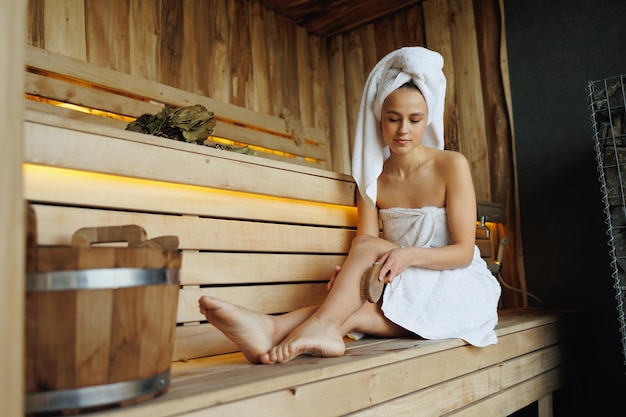 Foto schöne frau in weißem bademantel und handtuch auf dem kopf macht körpermassage mit trockener holzbürste mit natürlichen borsten in der sauna