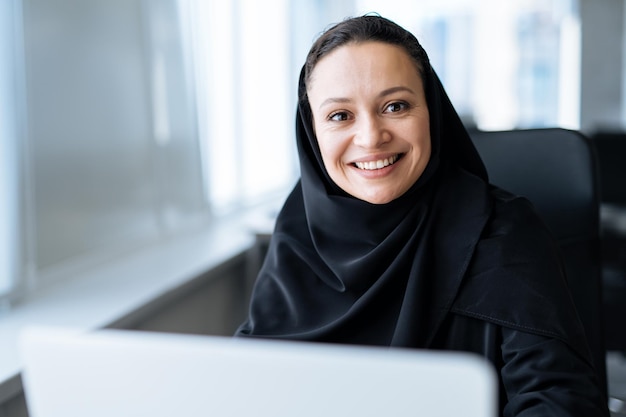 Schöne Frau mit Abaya-Kleid, die an ihrem Computer arbeitet. Angestellte mittleren Alters bei der Arbeit in einem Geschäftsbüro in Dubai. Konzept über Kulturen und Lebensstil des Nahen Ostens