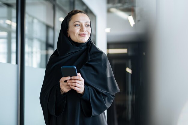 Schöne Frau mit Abaya-Kleid, die Dokumente bearbeitet und druckt. Angestellte mittleren Alters bei der Arbeit in einem Geschäftsbüro in Dubai. Konzept über Kulturen und Lebensstil des Nahen Ostens