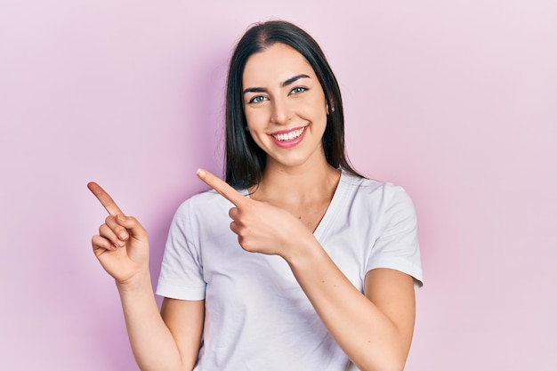 Schöne Frau mit blauen Augen, die lässiges weißes T-Shirt trägt, lächelt und in die Kamera schaut, die mit zwei Händen und Fingern zur Seite zeigt.