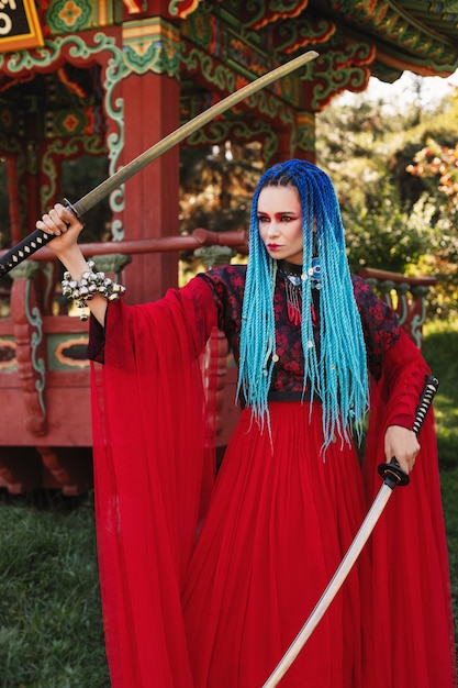 schöne Frau mit blauen Dreadlocks im roten transparenten Stoffkleid mit zwei Katanas