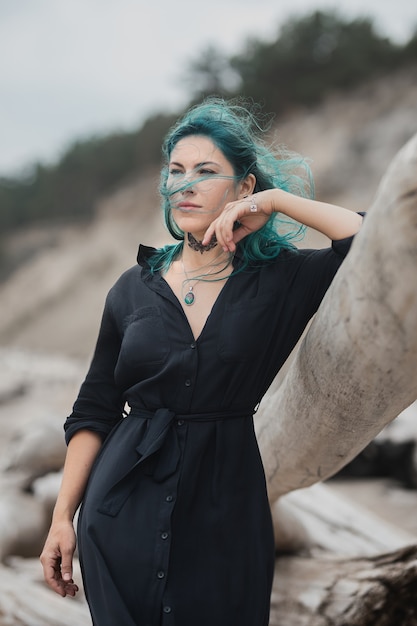 Schöne Frau mit blauen Haaren steht bei windigem Wetter am Strand und lehnt sich an einen Baumstamm