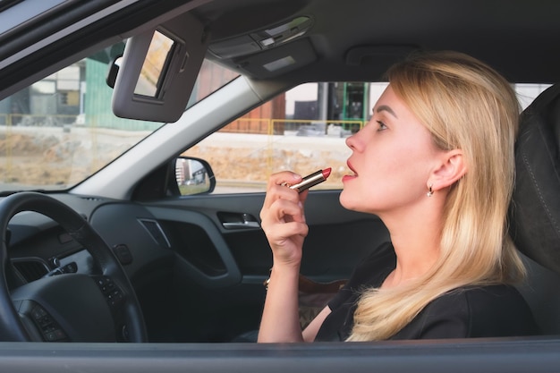 Schöne Frau mit blonden Haaren, die ein Auto fährt, malt ihre Lippen mit Lippenstift