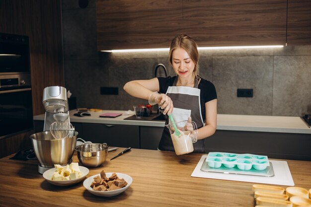 Schöne Frau mit blonden Haaren, gekleidet in eine Schürze, peitscht Sahne mit einem Mixer in einer modernen Küche