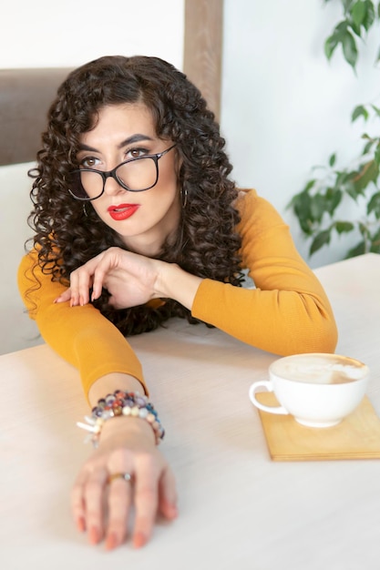 Schöne Frau mit Brille legte sich auf ihren Arm, während sie mit einer Tasse Kaffee an einem Tisch in einem Café saß