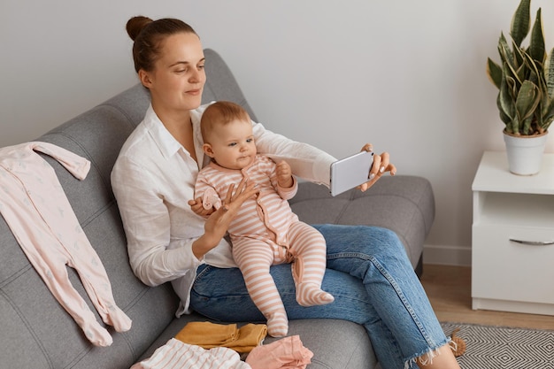 Schöne Frau mit dunklem Haar und Brötchenfrisur, die Freizeitkleidung trägt, die auf Husten sitzt, mit Kleinkindbaby mit Nippel in der Hand, Familie mit Livestream, Videoanruf oder Selfie.