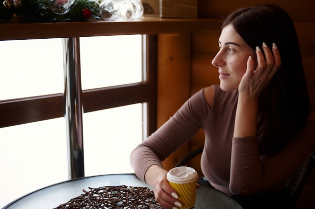 Schöne Frau mit einem Pappbecher des leckeren heißen Getränks, das am Café sitzt und aus dem Fenster schaut. Kaffeepausenzeit